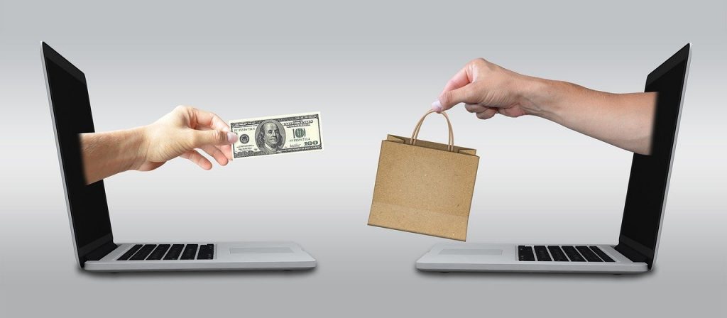 image of two laptop computers faceing each otehr with arms coming out of the screen, arm on the left has a one hundred dollar bill and is a female hand and the arm on the right is a male hand with a small brown bag with string type handles. Indicative of online selling and online buying of items. 