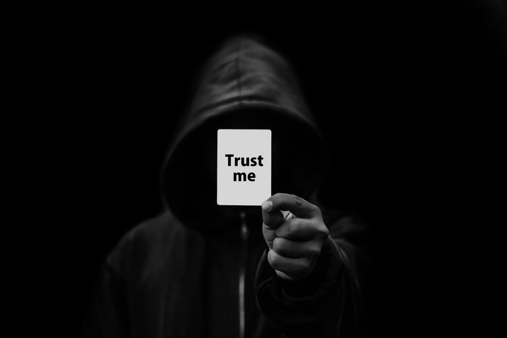 Image of a dark background with a man in a black hoodie holding a trust me card up indicated of shady charachters selling online.